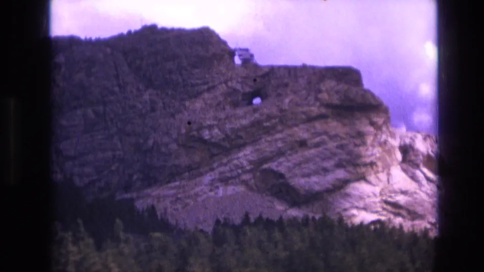 1974 8mm South Dakota frame showing a layered rocky formation with a small dark opening, evergreen forest below, purple-tinted sky, and visible dark borders from the film scan.