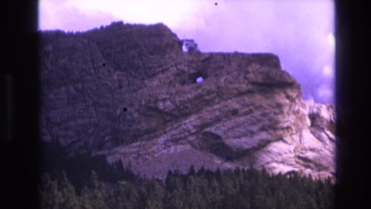 Layered rocky mountainside above a dark evergreen forest under a purple-tinted sky.