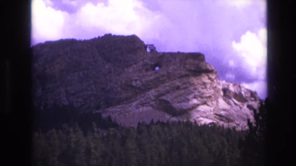1974 South Dakota 8mm frame showing a rugged rock slope above a dense treeline, with bright clouds and purple color shift.
