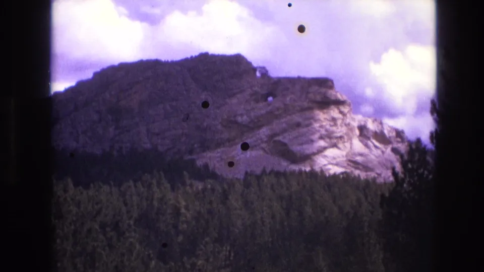 1974 South Dakota 8mm frame with a rugged ridge above forest, purple-tinted clouded sky, dark edge vignetting, and circular dust marks.