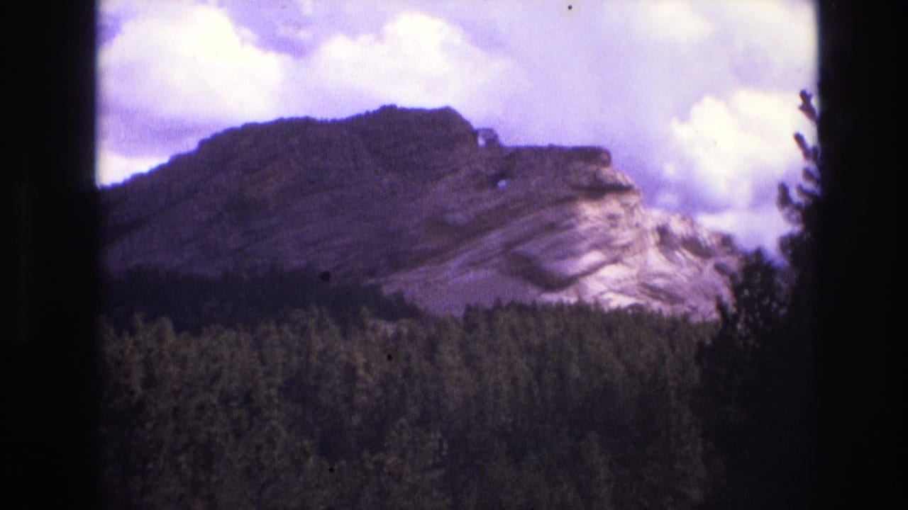 1974 8mm home-movie frame showing a rocky South Dakota mountain ridge rising above dark evergreen forest under bright, cloud-filled sky.