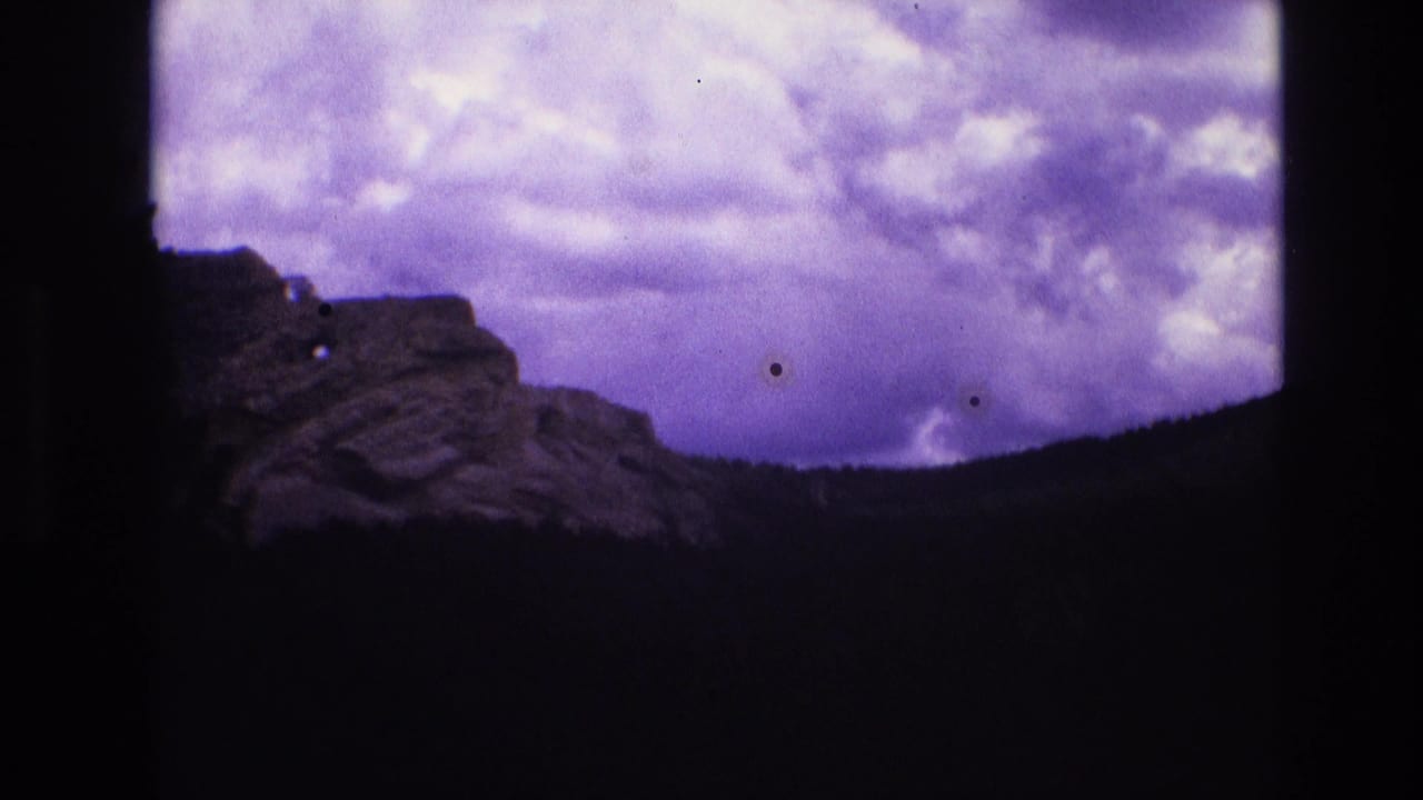 1974 8mm frame with a distant rocky formation partly centered above a dark forested slope and overcast purple-tinged sky.