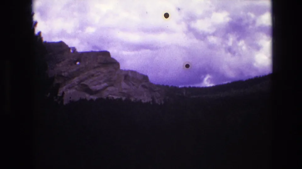 1974 8mm South Dakota landscape frame showing a rocky outcrop above shadowed evergreen forest with visible dust marks and cloudy sky.