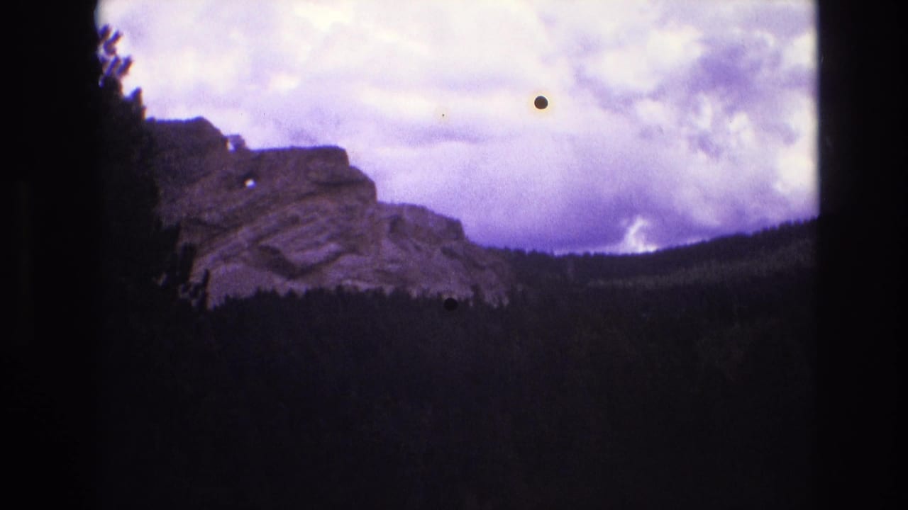 1974 South Dakota 8mm frame showing a rocky ridge and dark forested valley beneath a purple-shifted storm sky, with black film borders and dust specks visible.