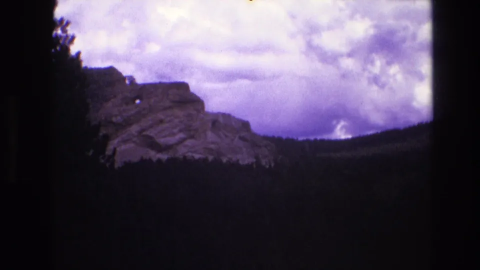 1974 South Dakota 8mm frame showing a closer rocky ridge, dark trees in the valley, and a bright cloud bank in a purple-tinted sky.