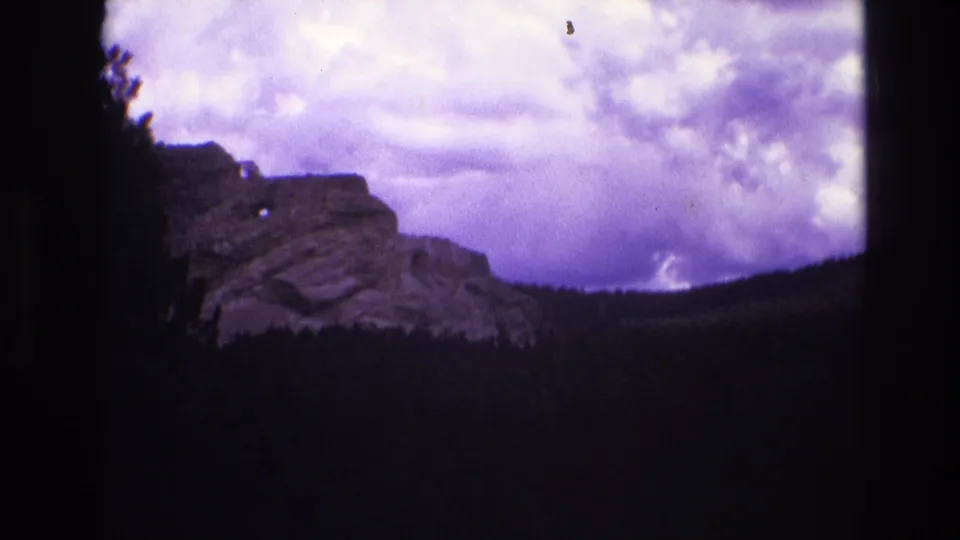 1974 South Dakota 8mm frame showing the rocky ridge and forested valley, with a small white horse statue or figure visible near the ridge under a cloudy purple sky.