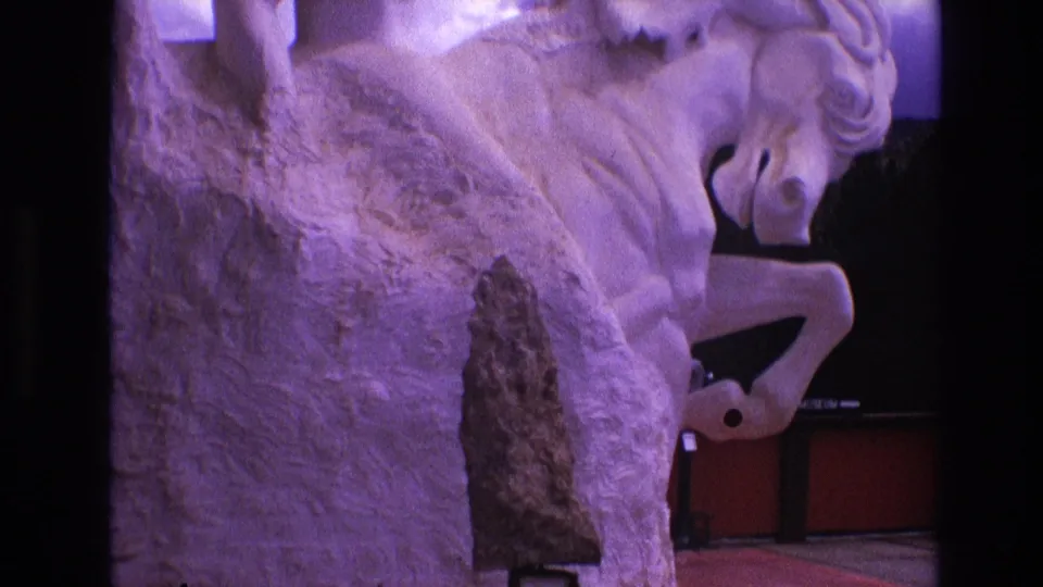 1974 South Dakota 8mm frame showing a close, slightly low view of a white rearing horse sculpture washed in purple light, with a dark pointed object mounted before it.