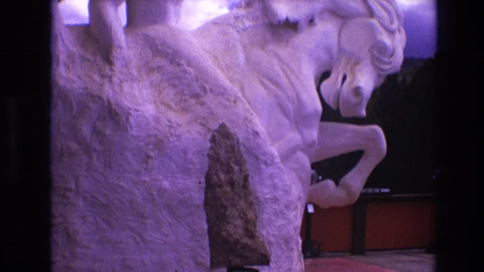 1974 South Dakota 8mm frame showing the rearing horse sculpture from a close visitor perspective, with purple light across the stone and a dark pointed mounted object in front.