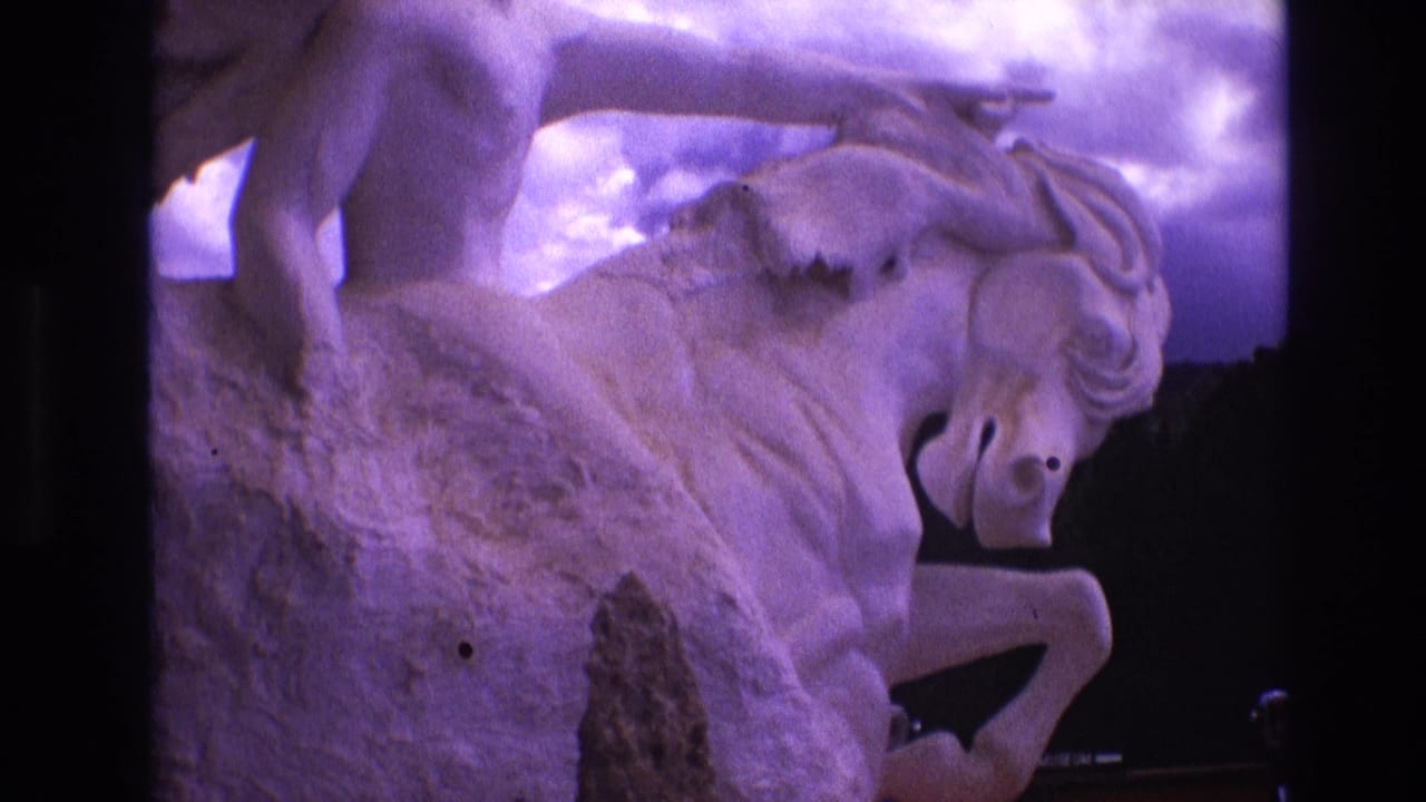 1974 South Dakota 8mm frame showing a close visitor view of a stone or concrete sculpture with a male rider above a rearing horse, tinted purple by the film image.