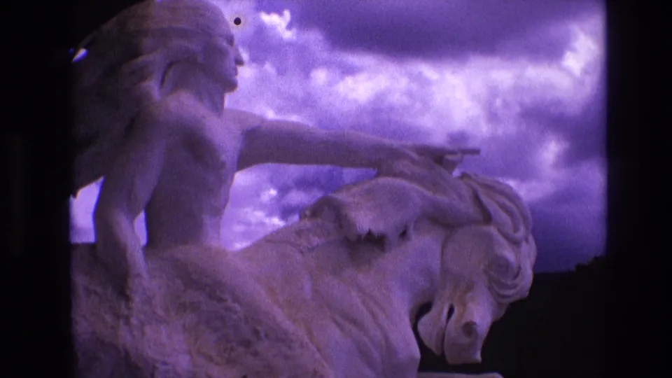 1974 South Dakota 8mm frame showing the horse-and-rider sculpture in close view, with the rider's arm crossing the cloudy sky and dark film borders visible at the edge.