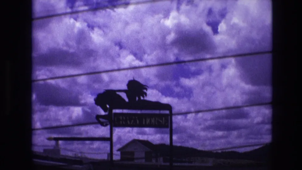 Purple-tinted 1974 South Dakota 8mm frame showing the CRAZY HORSE sign below overhead wires with buildings and hills in the distance.