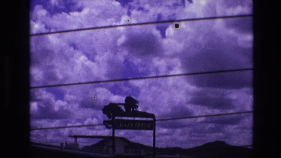 Final 1974 South Dakota frame with the CRAZY HORSE sign smaller in view, set against heavy clouds, utility lines, and low roadside structures.