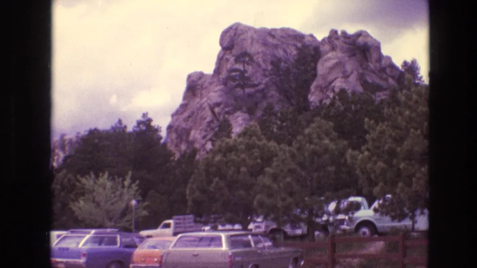A wide 1974 South Dakota home-movie view shows Mount Rushmore above trees, with parked 1970s cars and a truck in the foreground under a partly cloudy sky.
