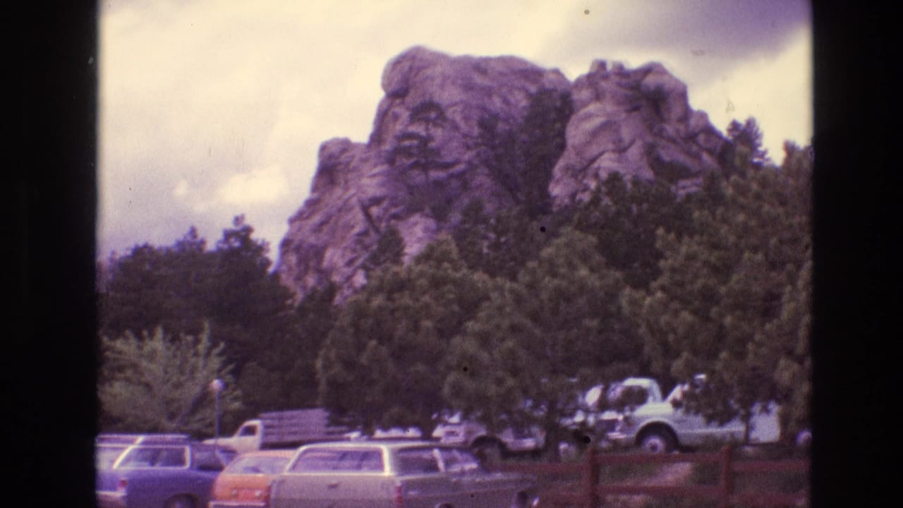 1974 South Dakota 8mm frame showing a rocky outcrop above pine trees, with parked 1970s cars and a light pickup truck in the foreground.