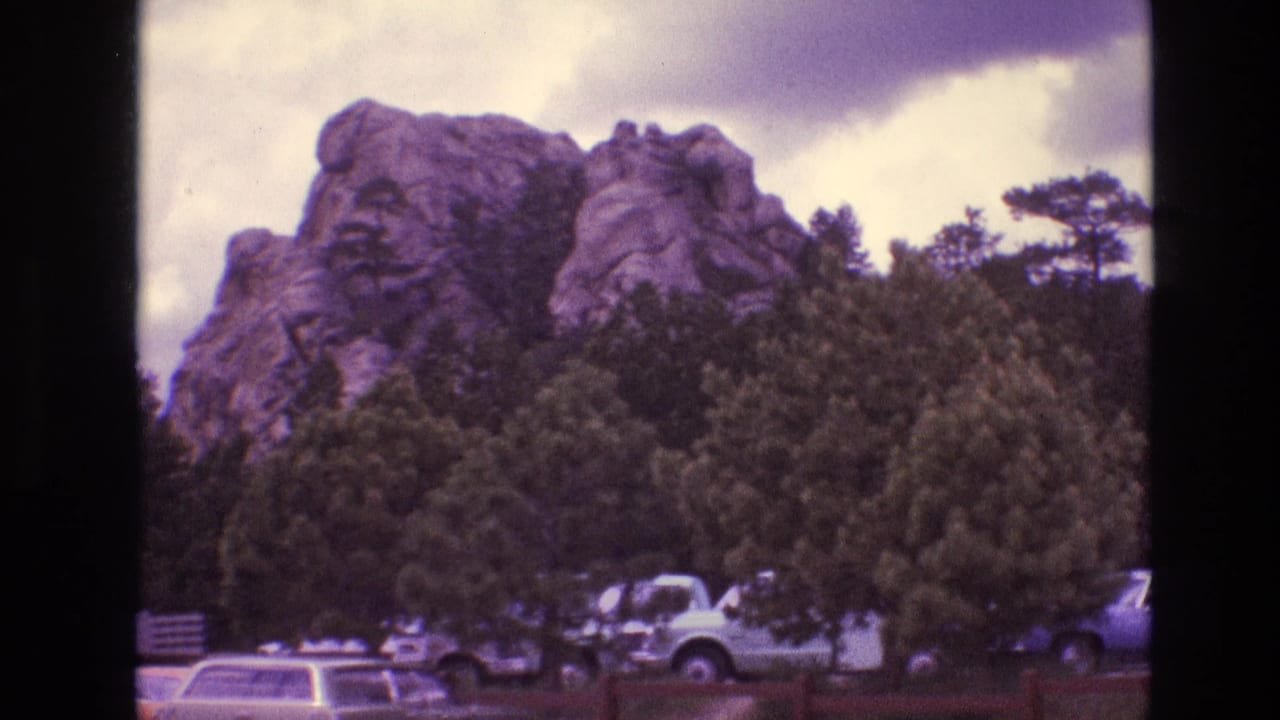 1974 South Dakota tourist-area view with a rugged rock formation rising behind trees and 1970s vehicles parked along the roadside.