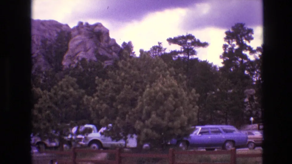 Handheld 8mm frame from South Dakota in 1974 showing trees and parked 1970s vehicles near a scenic rock formation.