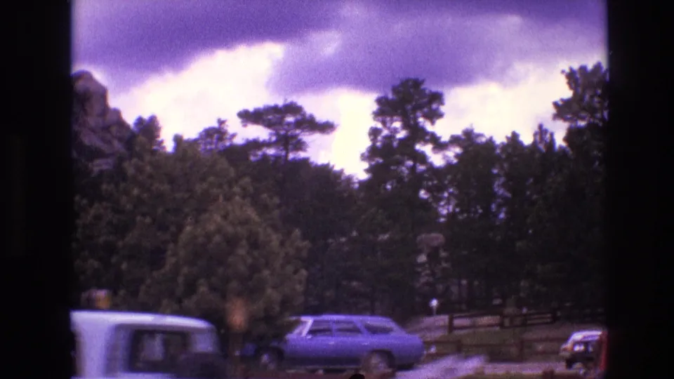 1974 South Dakota home-movie frame with a parked blue car, roadside fencing, dense trees, and partial rock formations under an overcast sky.