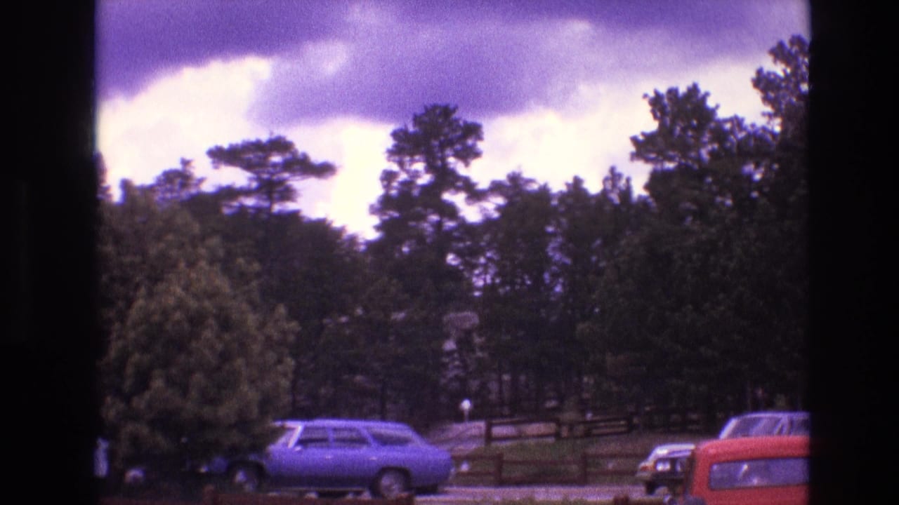 1974 South Dakota 8mm frame showing parked 1970s cars near pine trees and a wooden fence beneath a cloudy sky.