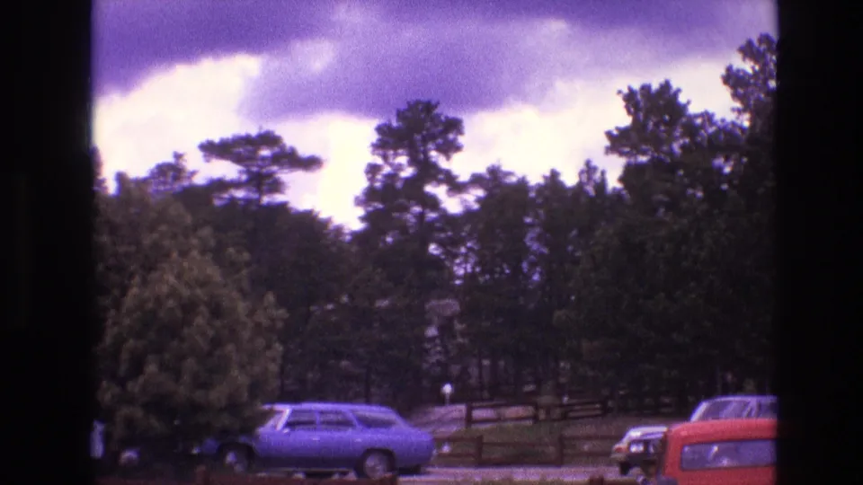 1974 South Dakota 8mm frame showing parked 1970s cars near pine trees and a wooden fence beneath a cloudy sky.