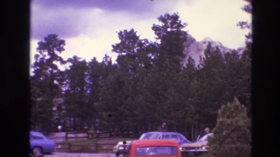 1974 South Dakota home-movie frame with parked cars, distant people, pine trees, and a rocky mountain partly visible beyond the treeline.
