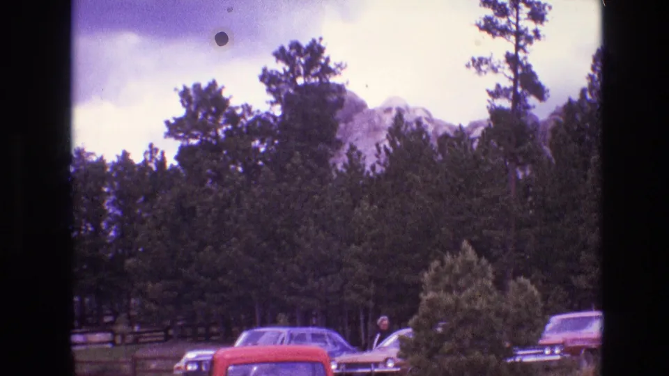 1974 South Dakota 8mm frame showing a rocky mountain behind dense pine trees, with parked cars and a distant person near the lower edge.