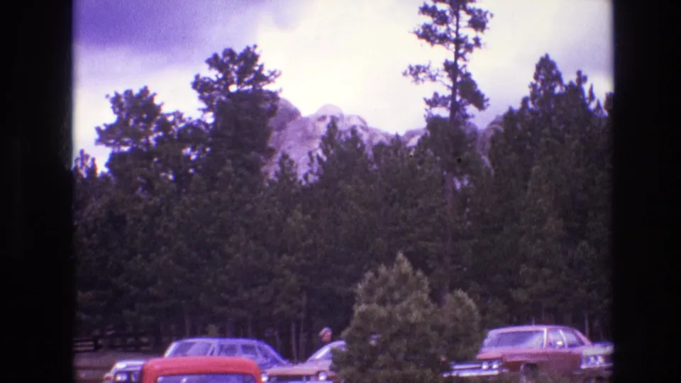 1974 South Dakota home-movie frame showing pine trees and a rocky mountain beyond a parking area with several 1970s cars and a distant person near an open car door.