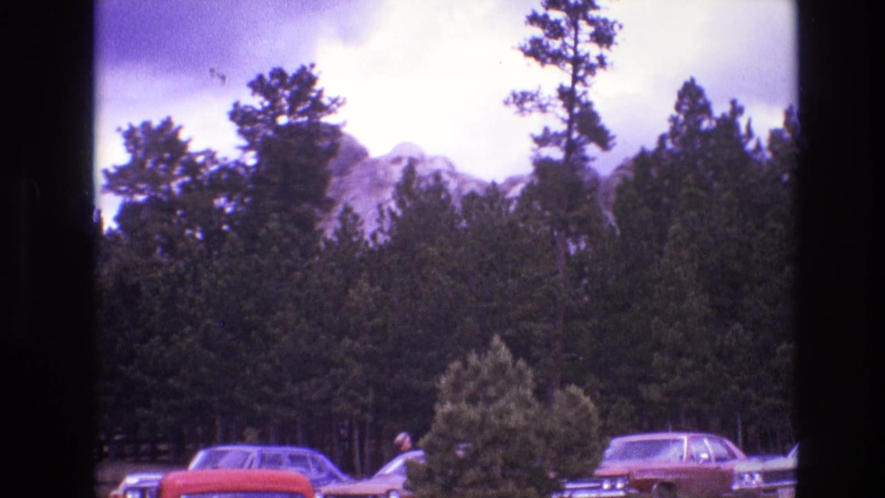 1974 South Dakota 8mm frame showing Mount Rushmore partly obscured by pine trees, with classic parked cars at the bottom of the view.