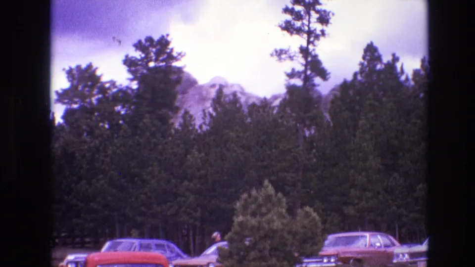 1974 South Dakota 8mm frame showing Mount Rushmore partly obscured by pine trees, with classic parked cars at the bottom of the view.