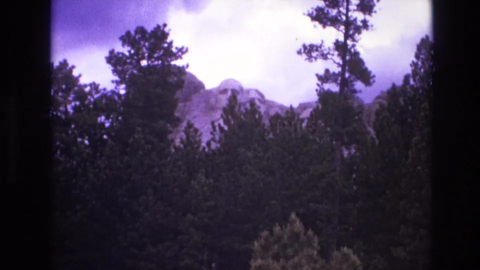 1974 South Dakota home-movie frame with Mount Rushmore visible behind dense pine trees and a bright overcast sky.