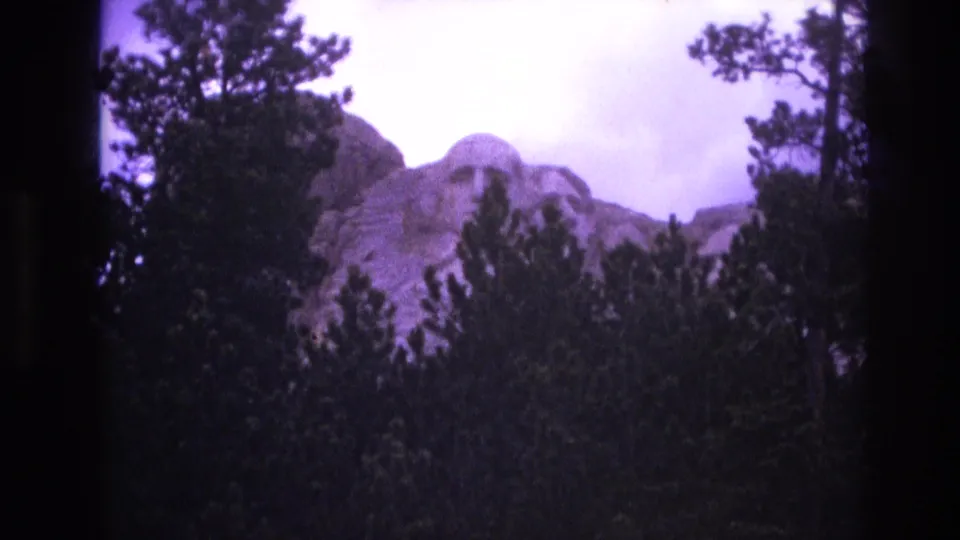1974 South Dakota 8mm frame showing the presidential carvings of Mount Rushmore rising behind a dark line of pine trees.
