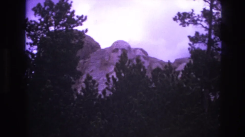 1974 South Dakota home-movie frame of Mount Rushmore framed by trees, with soft focus, grain, and a purple-tinted cloudy sky.