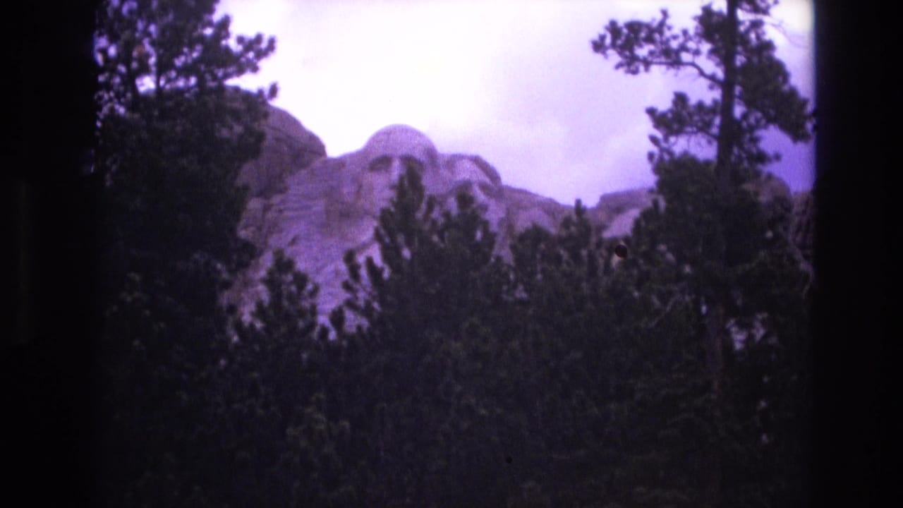1974 8mm home-movie frame showing Mount Rushmore in South Dakota partly obscured by pine trees, with a purple color cast and dark edge vignetting.
