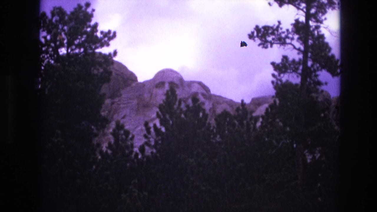 1974 South Dakota home-movie frame with Mount Rushmore visible through trees, softened by 8mm grain, darkened edges, and aged purple film color.