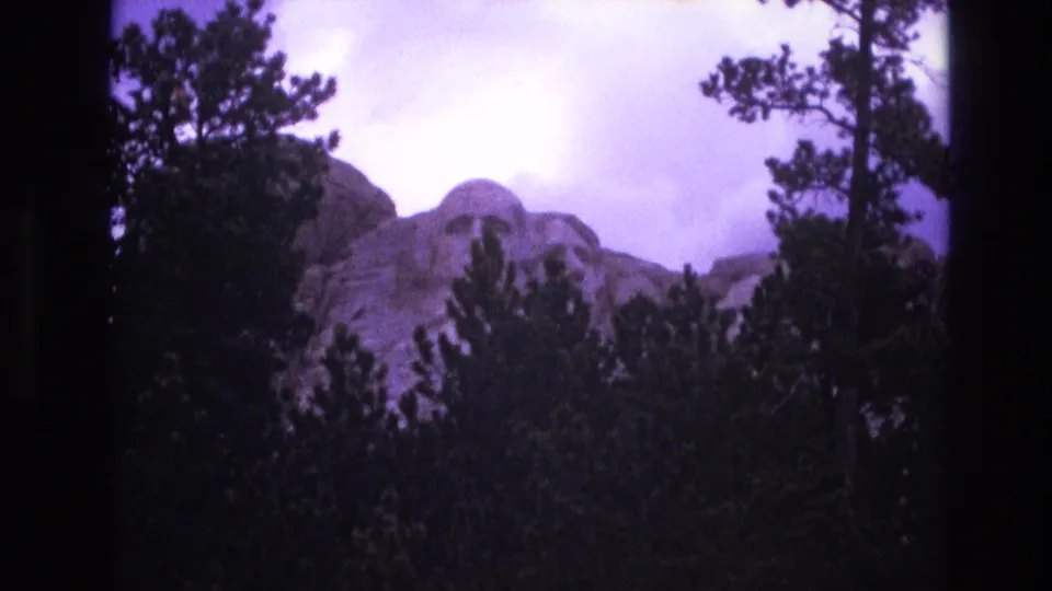 1974 8mm frame from South Dakota showing Mount Rushmore behind dense pine trees, with visible grain, soft focus, and dark home-movie framing.
