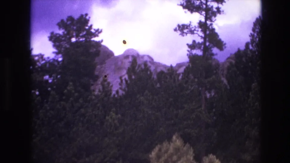 1974 South Dakota 8mm frame showing the Mount Rushmore faces through a wooded foreground, with purple film aging, dust spots, and slight motion softness.