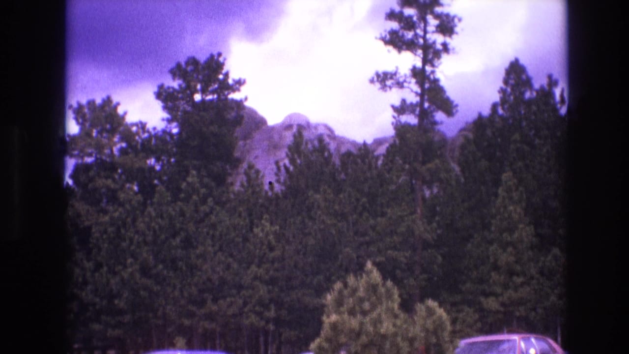 1974 South Dakota 8mm frame showing pine trees, distant hills, bright sky, and part of an older parked car near a scenic park path.