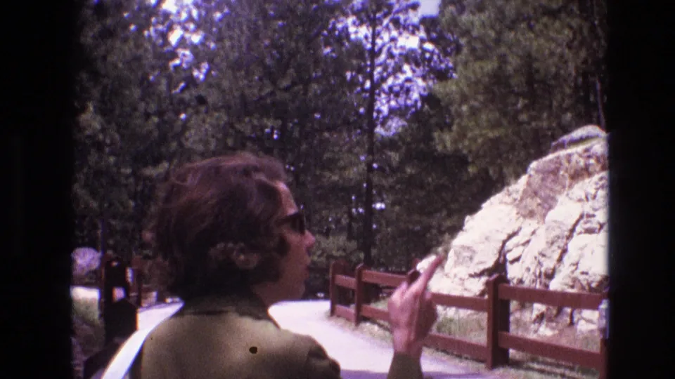 Young woman with curly hair, sunglasses, green jacket, and white purse walking along a 1974 South Dakota park path beside a wooden fence and rock formation.