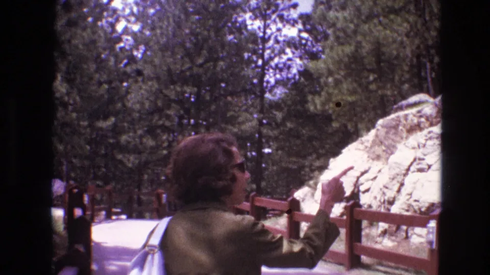1974 South Dakota home-movie frame of a young woman with sunglasses and white purse pointing toward a pale rock formation along a fenced park path.