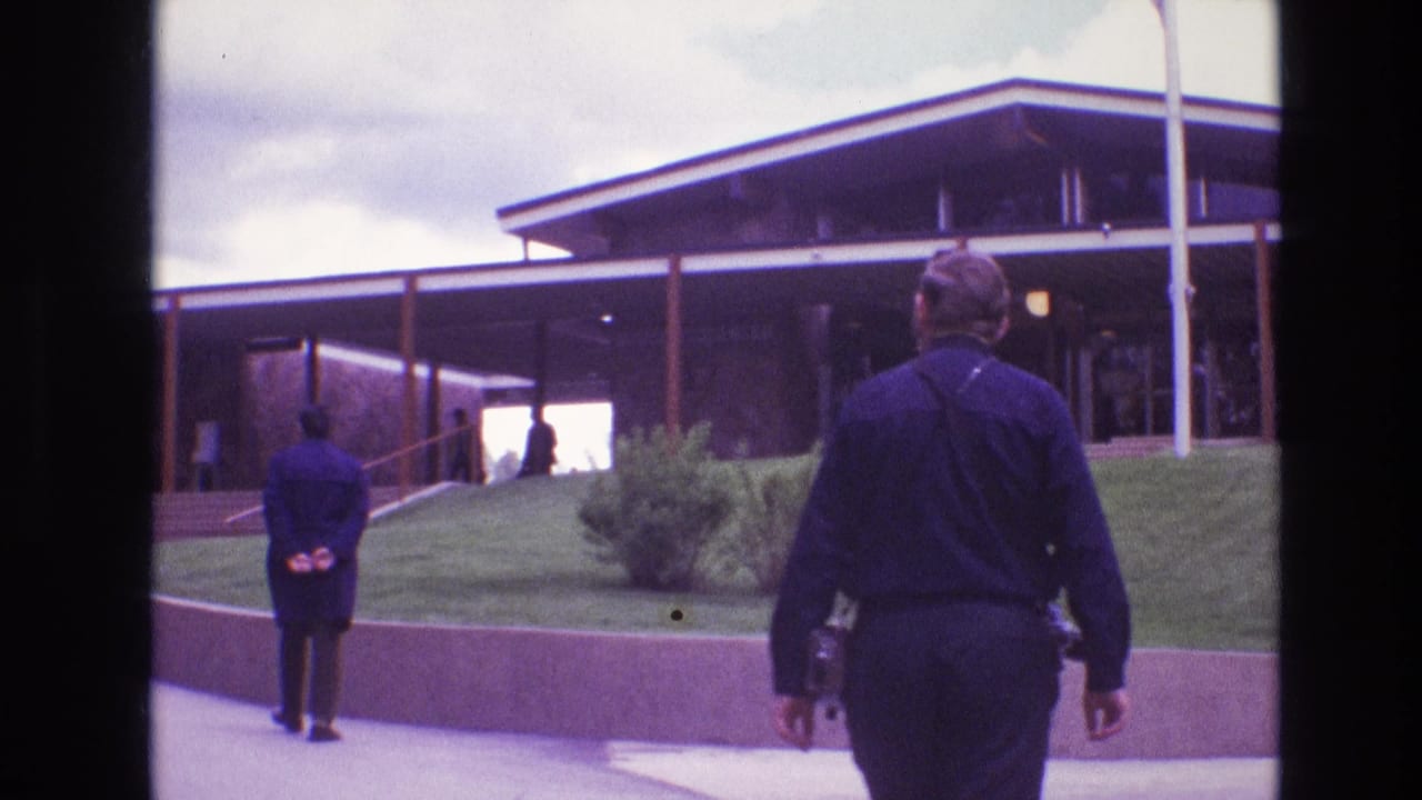1974 South Dakota 8mm frame showing two adults near a large flat-roofed modern building, with one man walking away from camera and another standing near the walkway.