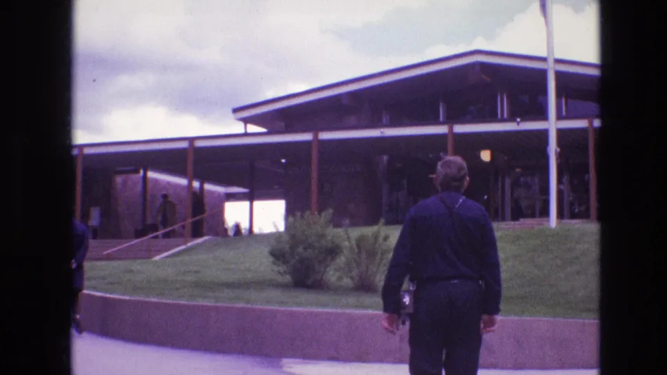 1974 South Dakota 8mm frame showing a man seen from behind on a paved approach to a mid-century modern building with lawn, retaining wall, and shaded entrance.