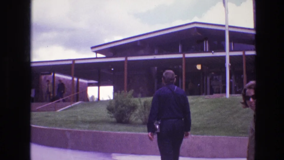 1974 South Dakota frame with a man walking toward a large modern building and a woman in sunglasses partly visible at the right edge.