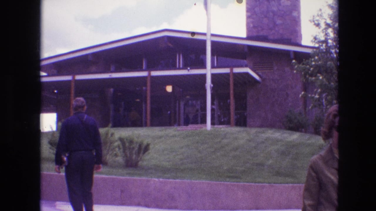 1974 South Dakota home-movie frame showing visitors approaching a low flat-roofed visitor-center building near Mount Rushmore, with flagpoles, lawn, shrubs, and visible 8mm frame borders.