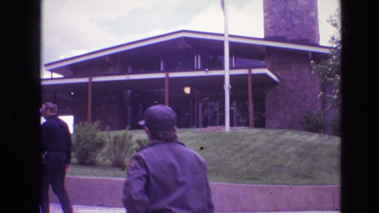 Tourists walk past a low visitor building with flagpoles and a grassy slope.