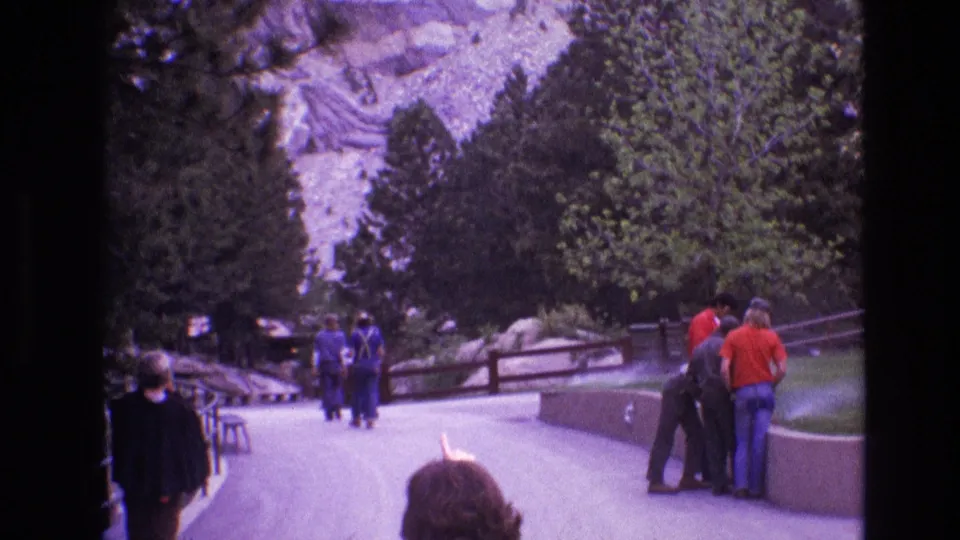 1974 Mount Rushmore home-movie frame showing tourists on a paved walkway, evergreen trees, railings, rocky cliff faces, and part of the presidential carvings high in the background.