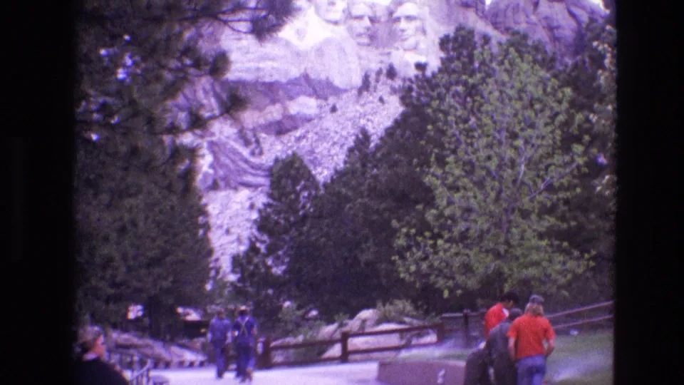 1974 South Dakota frame showing visitors near a railing at Mount Rushmore, with evergreen trees, rocky slope, and the presidential carvings visible in the background.