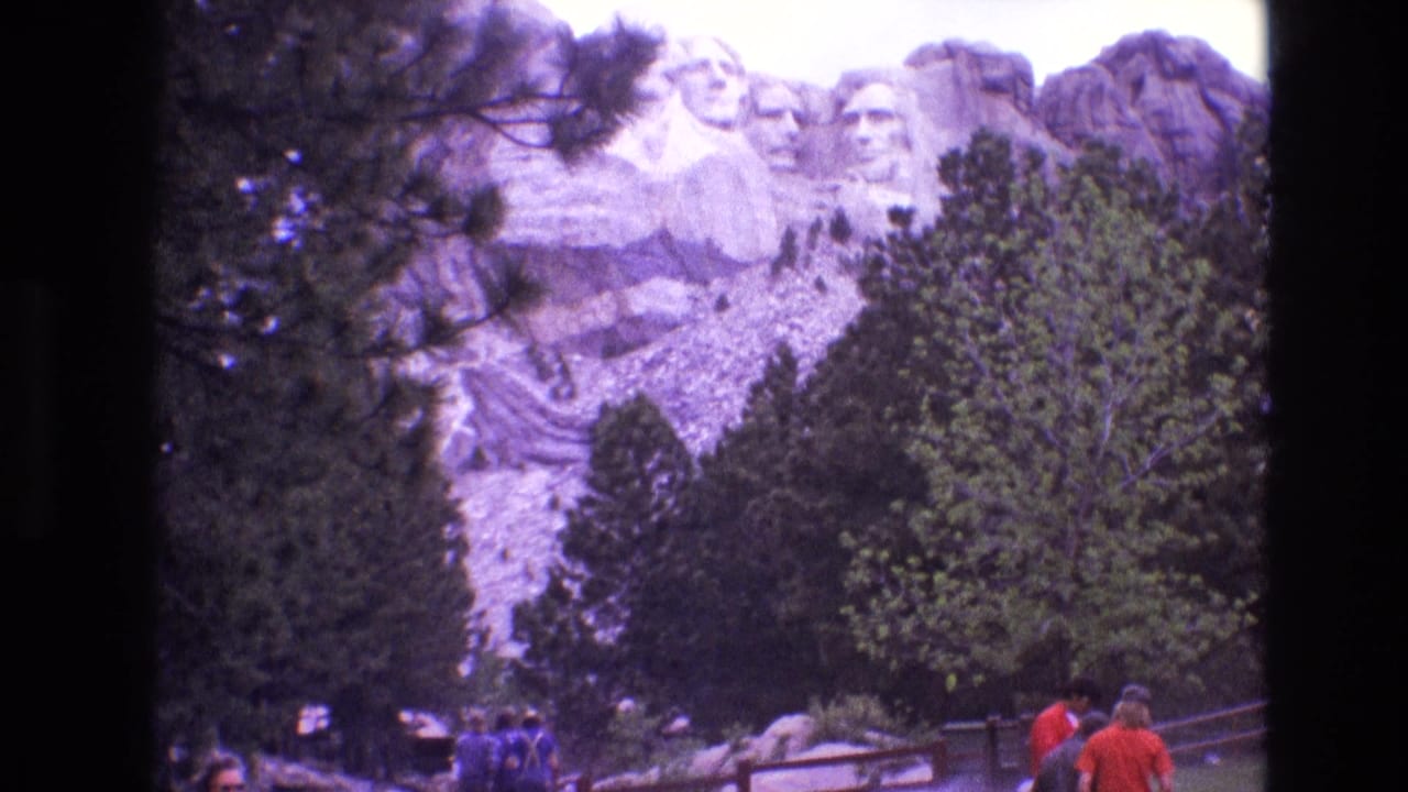 Mount Rushmore seen through trees with visitors on a pathway below.