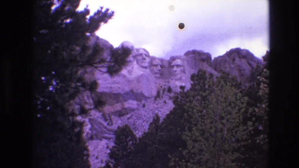 A handheld 8mm view shows Mount Rushmore above pine trees and rocky terrain, with aged purple color cast consistent with the supplied 1974 home-movie metadata.