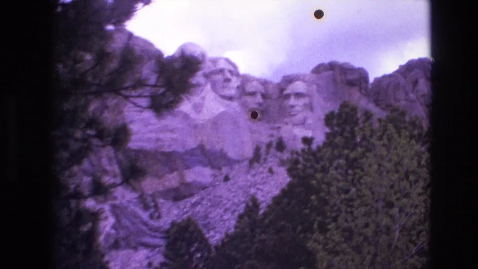 The final 1974 8mm frame holds on Mount Rushmore behind pine trees, with rocky slopes, cloudy sky, and visible film dust marks in the image area.