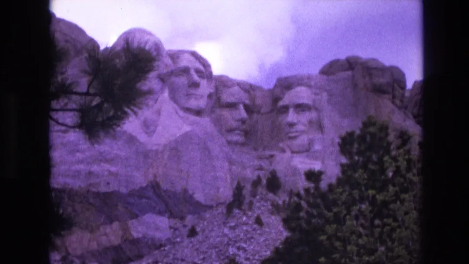 1974 South Dakota travel frame showing the Mount Rushmore presidential carvings above a rocky slope, with pine foliage along the lower edge.