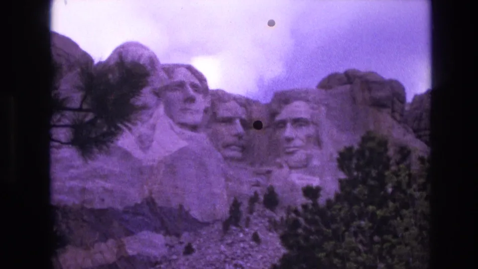 1974 8mm frame of Mount Rushmore with a closer visitor-shot view of the carved faces, visible purple color cast, dust marks, and trees at the bottom.